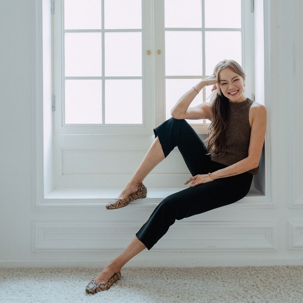 Jade sits on a white window sill in a bright room, wearing a brown sleeveless mock-neck top, black cropped pants, and brown snakeskin-patterned flat shoes.