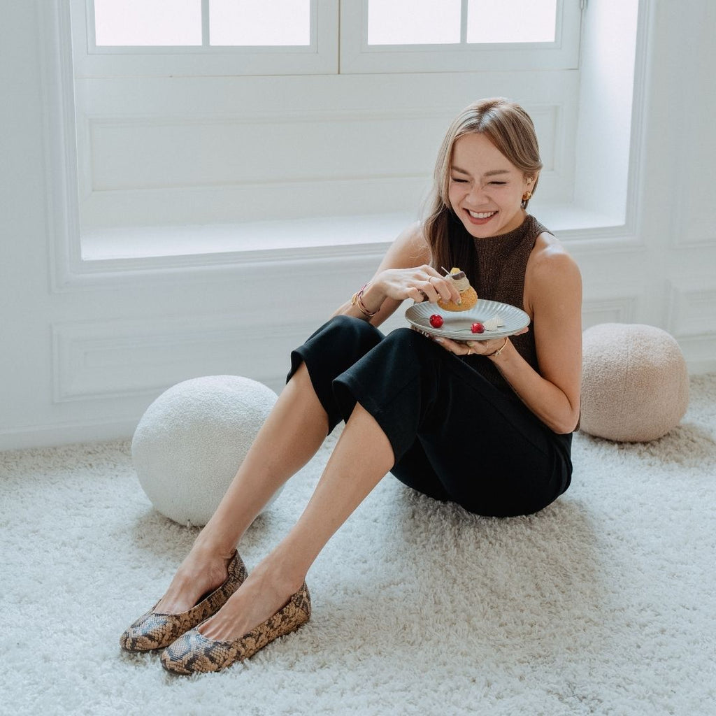 Jade sits on a white shag rug in a bright room, holding a plate with a dessert. She is wearing a dark brown sleeveless top, black wide-leg pants, and Embossed Python Prints Low Wedge Flats - Nude