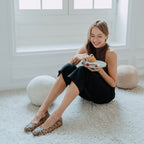Jade sits on a white shag rug in a bright room, holding a plate with a dessert. She is wearing a dark brown sleeveless top, black wide-leg pants, and Embossed Python Prints Low Wedge Flats - Nude